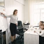 A female teacher guiding two girls during a lesson in a bright classroom with a whiteboard. — Professional Online Spanish Cla
